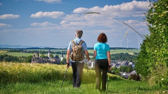 Wandern bei Kastellaun Zwei Wanderer gehen auf einem Wiesenweg mit Blick auf Kastellaun und Windräder.