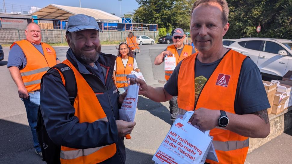 Markus Friedel (rechts) bietet einem Mitarbeiter von Schütz vor den Werkstoren eine Tüte zur Aktion für mehr Tarifverträge an.