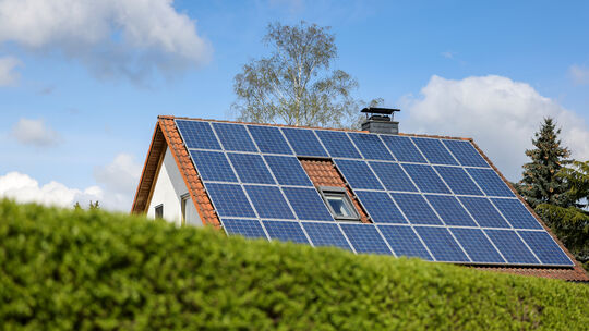 Ein Einfamilienhaus mit Solarpaneelen auf dem Hausdach steht hinter einer grünen Hecke. Immer mehr Unternehmen und Privathaushal Ein Einfamilienhaus mit Solarpaneelen auf dem Hausdach steht hinter einer grünen Hecke. Immer mehr Unternehmen und Privathaushal