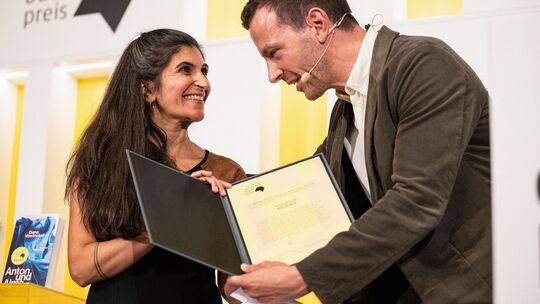 Die Gewinnerin des Deutschen Buchpreises 2026, Shida Bazyar erhält die Urkunde aus den Händen von Sebastian Guggolz, dem Vorsteh Die Gewinnerin des Deutschen Buchpreises 2026, Shida Bazyar erhält die Urkunde aus den Händen von Sebastian Guggolz, dem Vorsteh